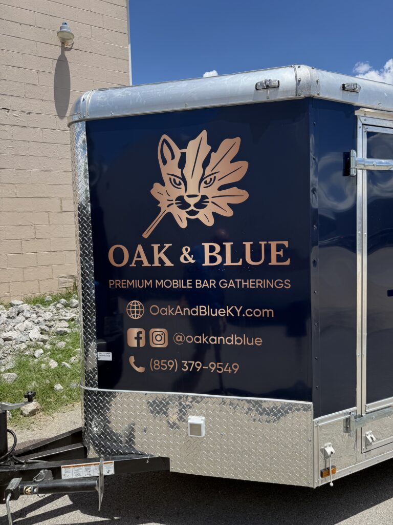 Front corner view of an Oak & Blue enclosed trailer wrap displaying logo, website, social media information, and phone number.